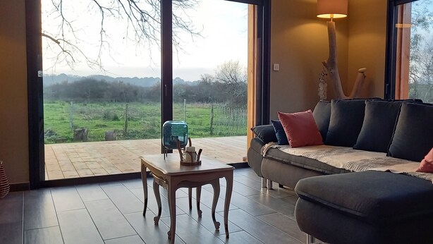 A modern living room with a large window, a sofa with multiple cushions, a small table with a teapot, and a decorative wooden piece.