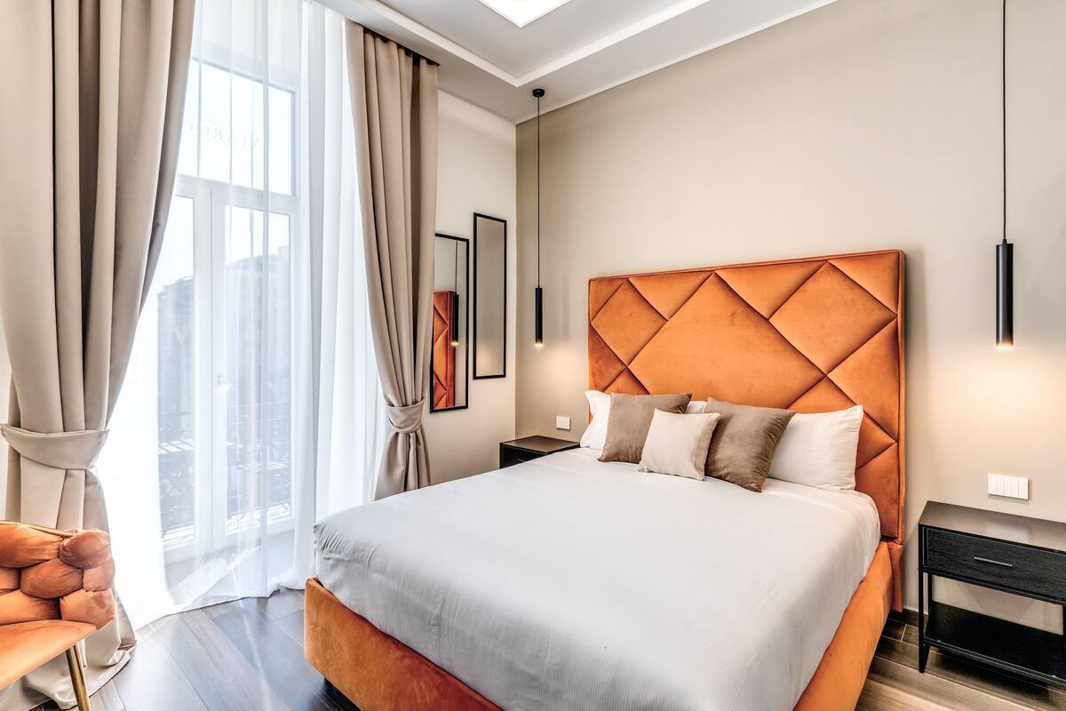 A modern hotel room with a large bed, a nightstand, a chair, and a window with curtains.