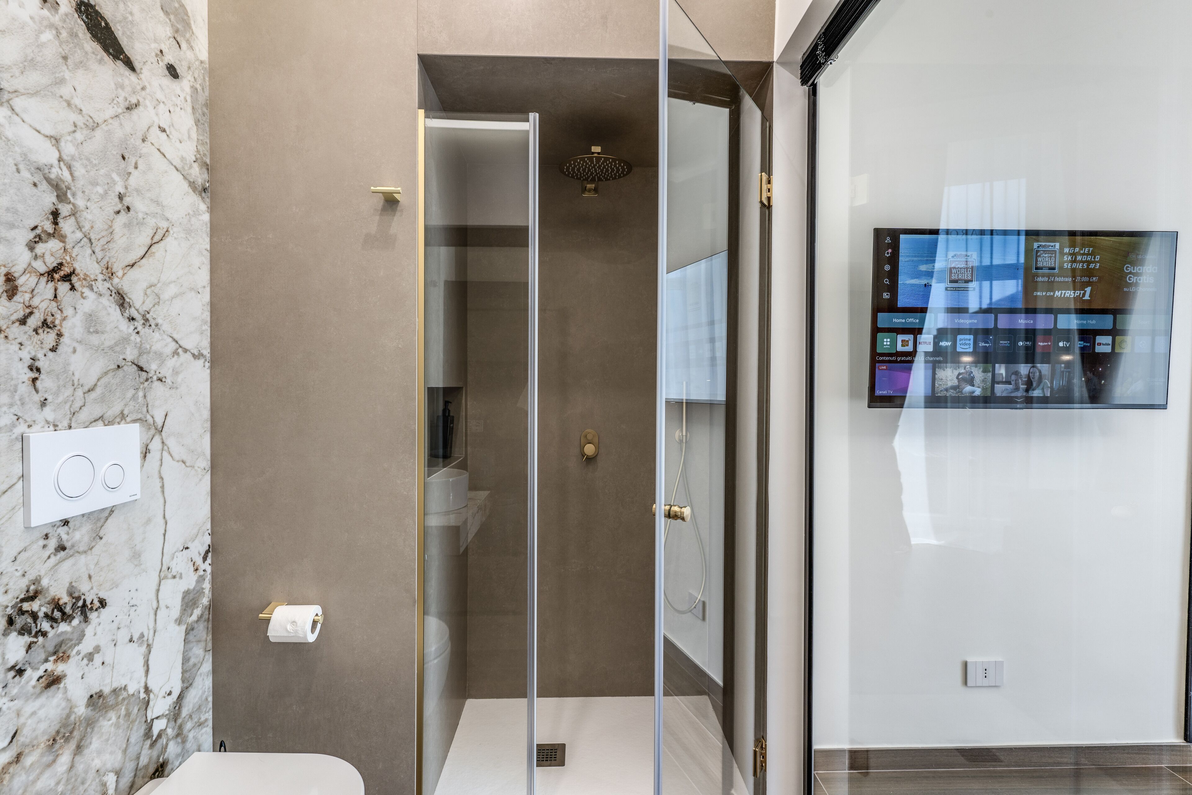 Un bagno moderno con box doccia in vetro, parete in marmo e schermo digitale che mostra informazioni dell'hotel.