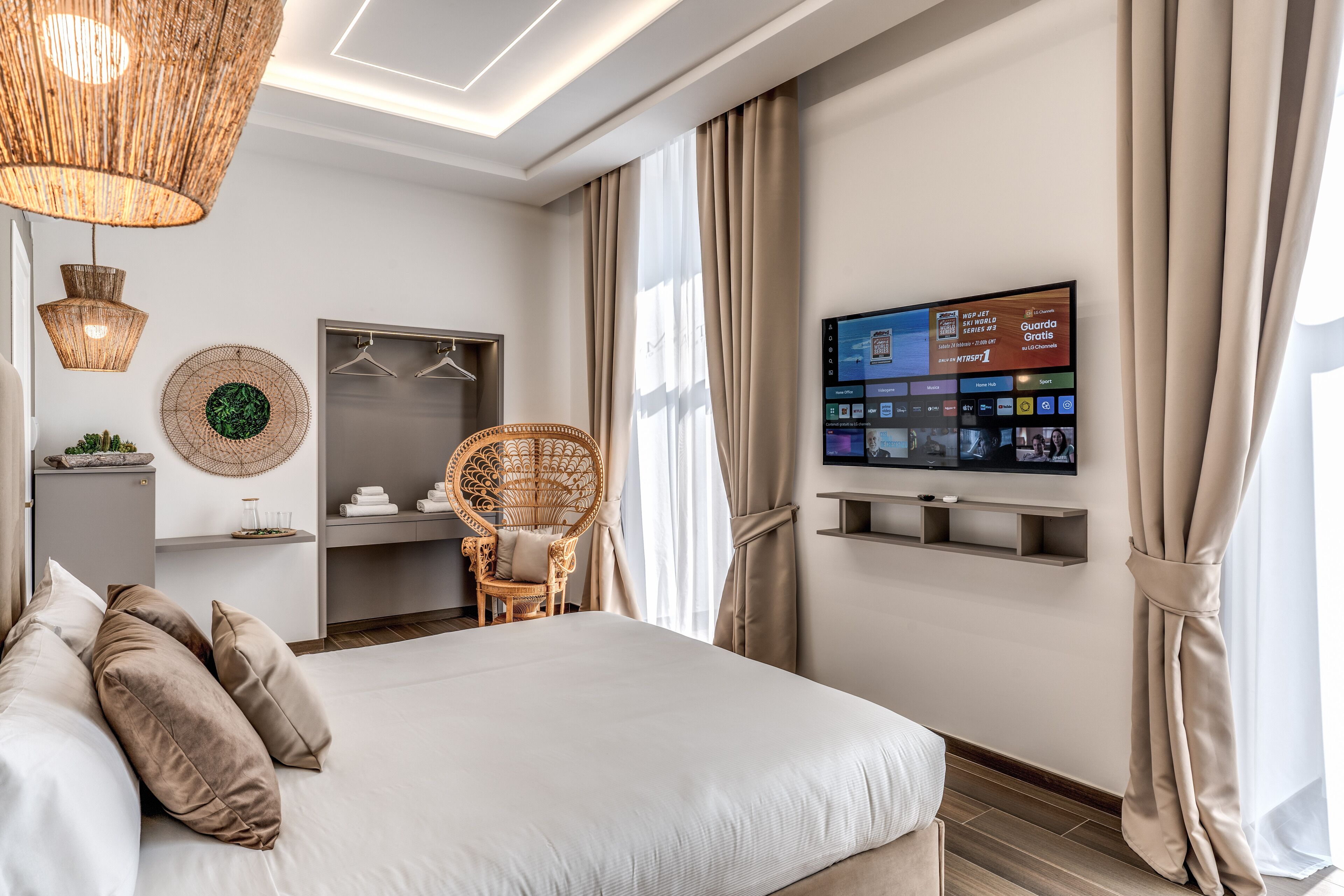 A modern hotel room with a large bed, a television mounted on the wall, a wardrobe, and a wicker chair.