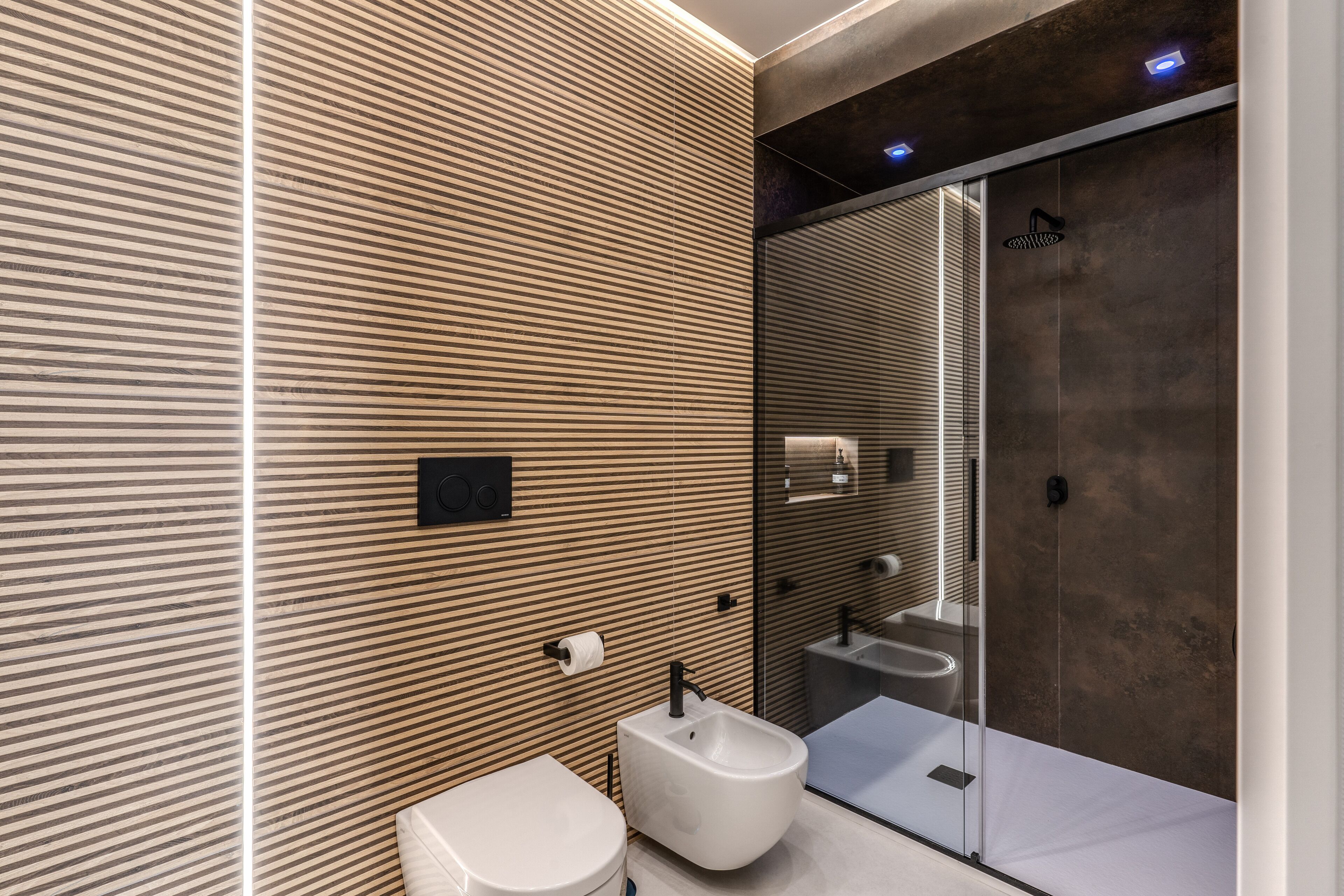 A modern bathroom with a wooden panel wall, a bidet, a toilet, and a glass-enclosed shower area.