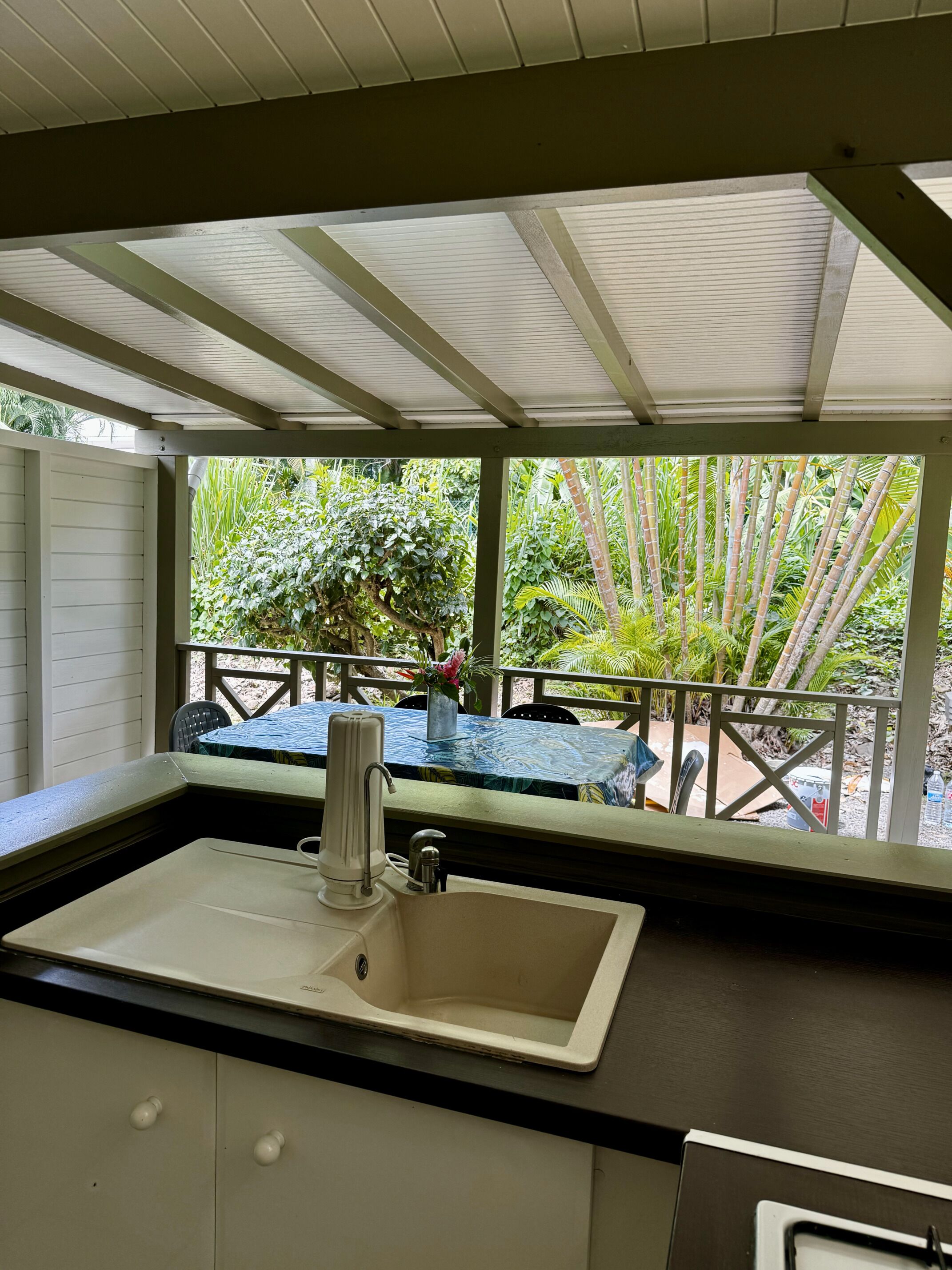 A covered outdoor kitchen area with a sink and a table, surrounded by lush greenery.