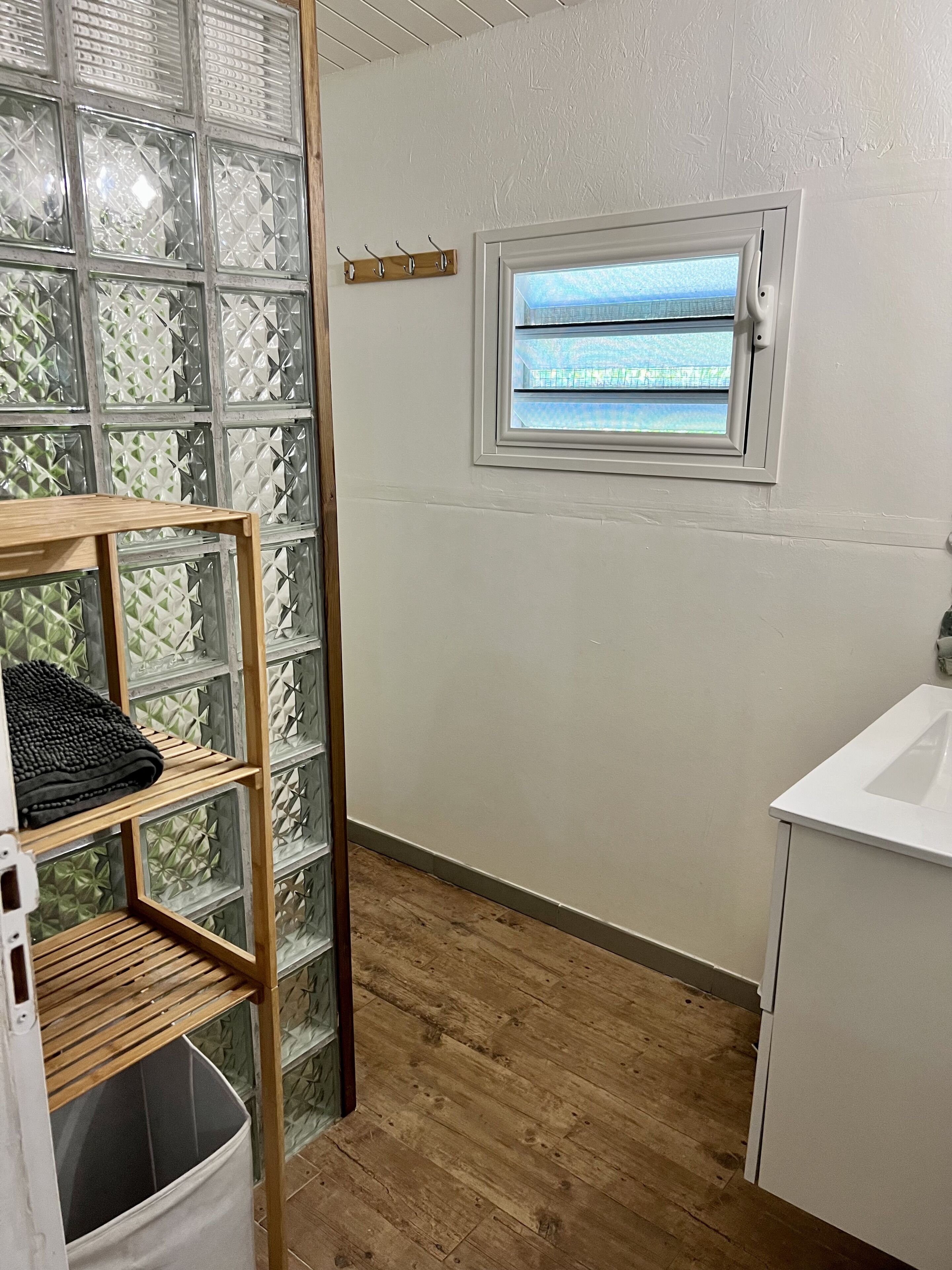 Een kleine, schone kamer met een houten vloer, een raam met uitzicht, een wit wastafelmeubel en een plankje met een handdoek.