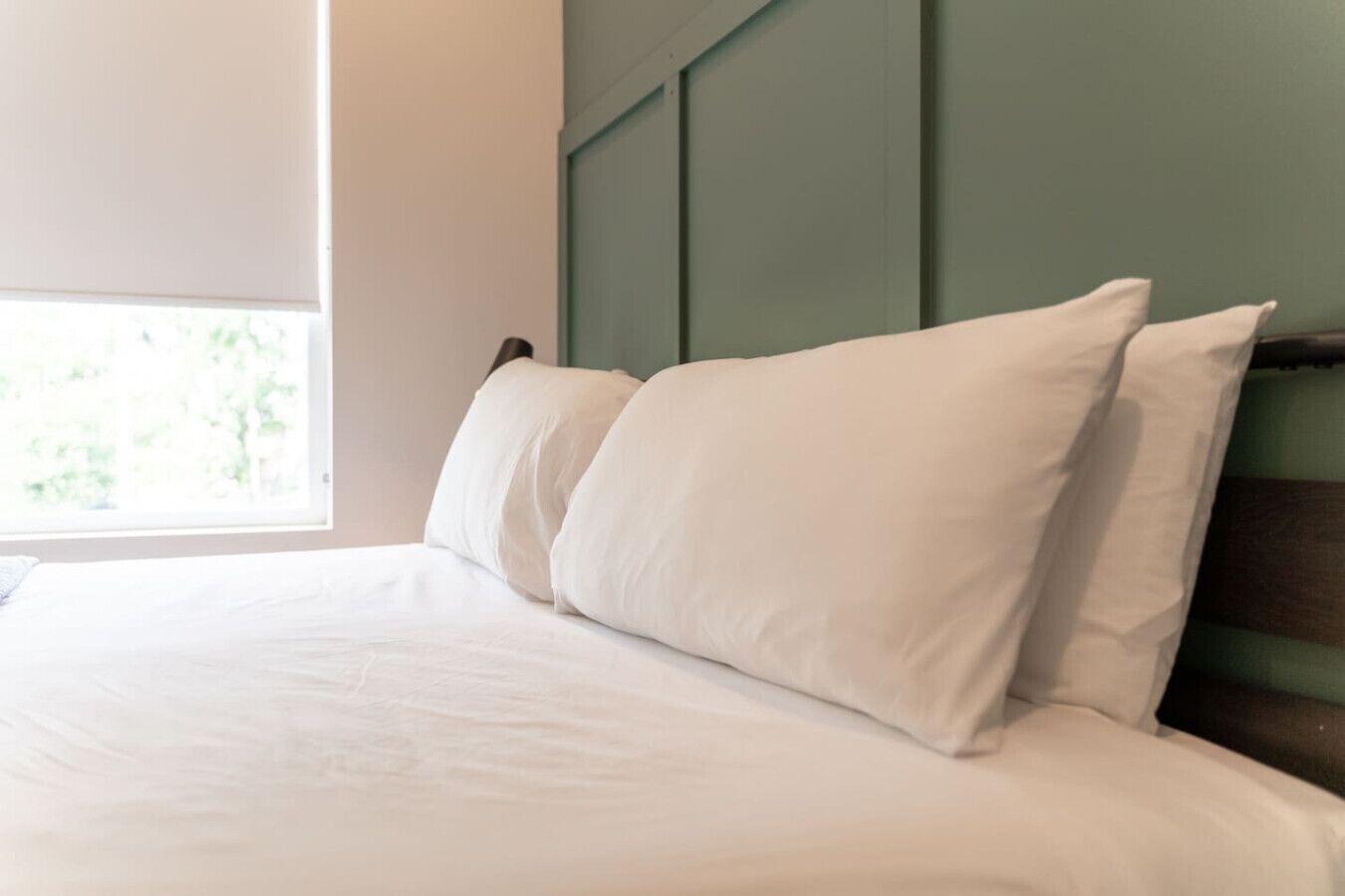 A neatly made bed with white linens and pillows, a wooden headboard, and a window with a view of greenery.