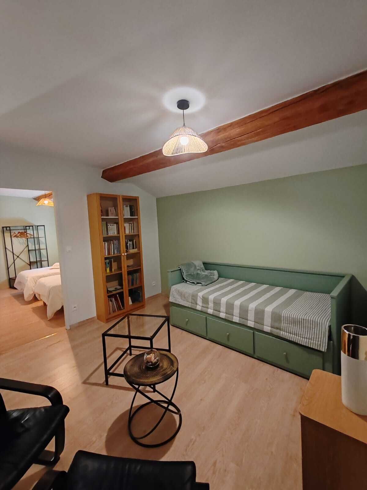 Una habitación pequeña y acogedora con un sofá verde, una estantería de madera, una mesa redonda y una cama al fondo.