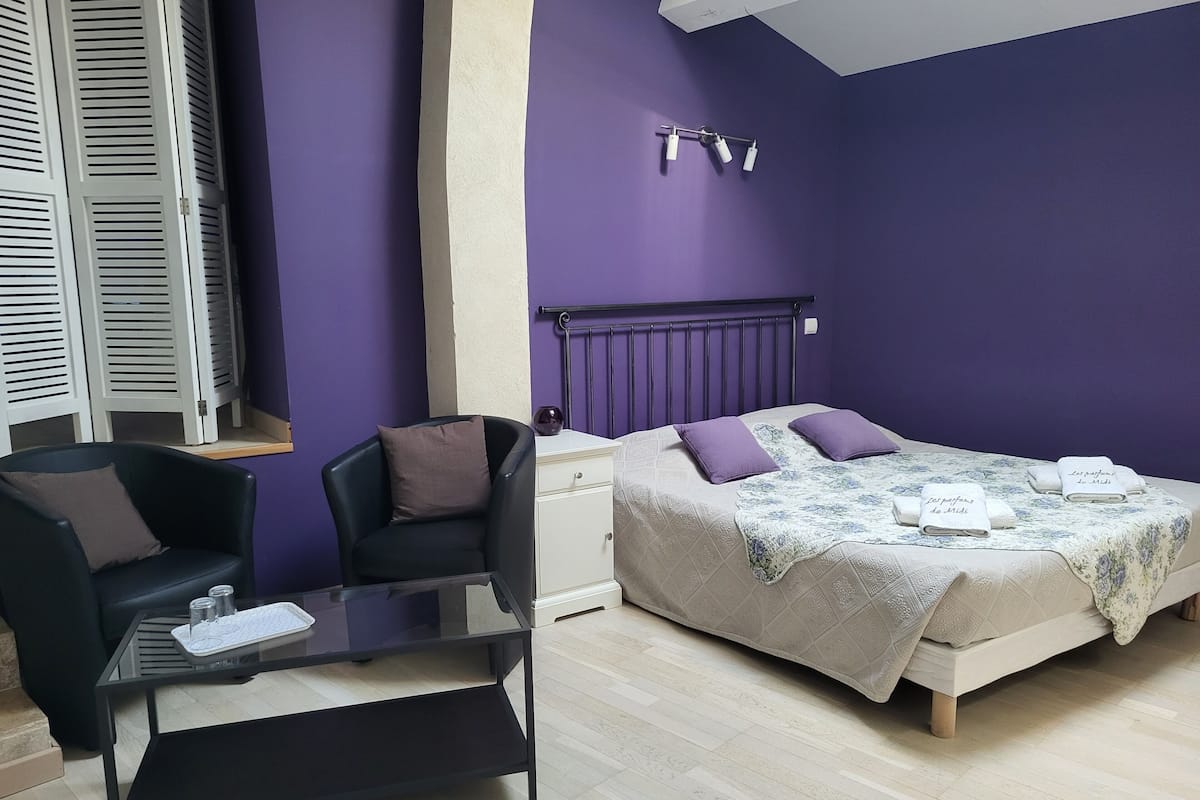 Un dormitorio con una cama, una silla, una mesita y una ventana con contraventanas.