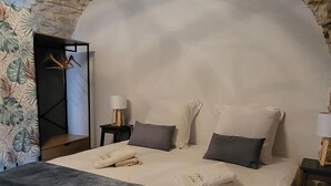 Un dormitorio con techo de piedra, una cama grande con ropa de cama blanca y gris, una mesita de noche de madera con una lámpara, y una pequeña mesa con una silla.