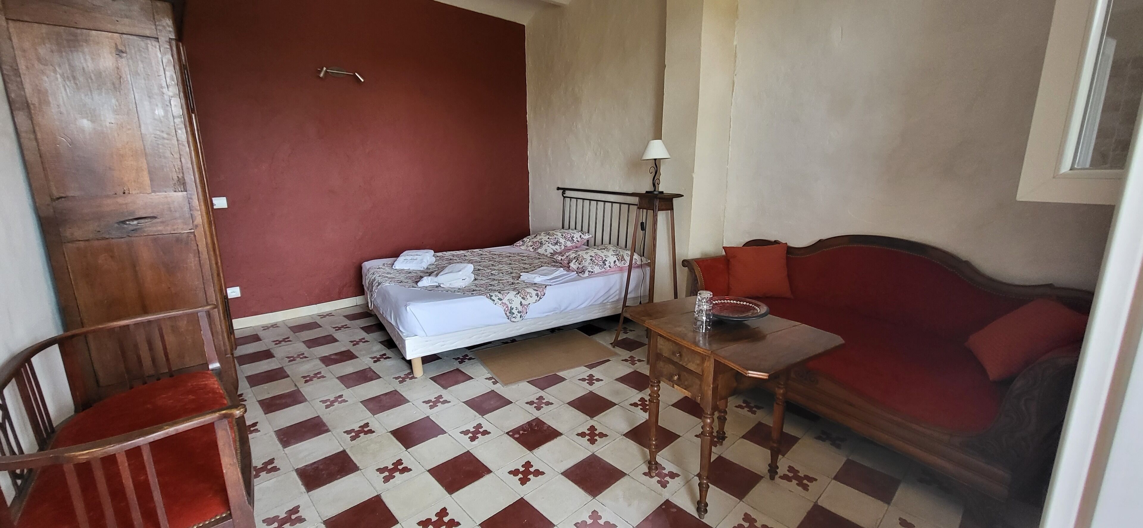 Una habitación pequeña con una cama, una silla de madera, una mesita con lámpara y un sofá rojo.