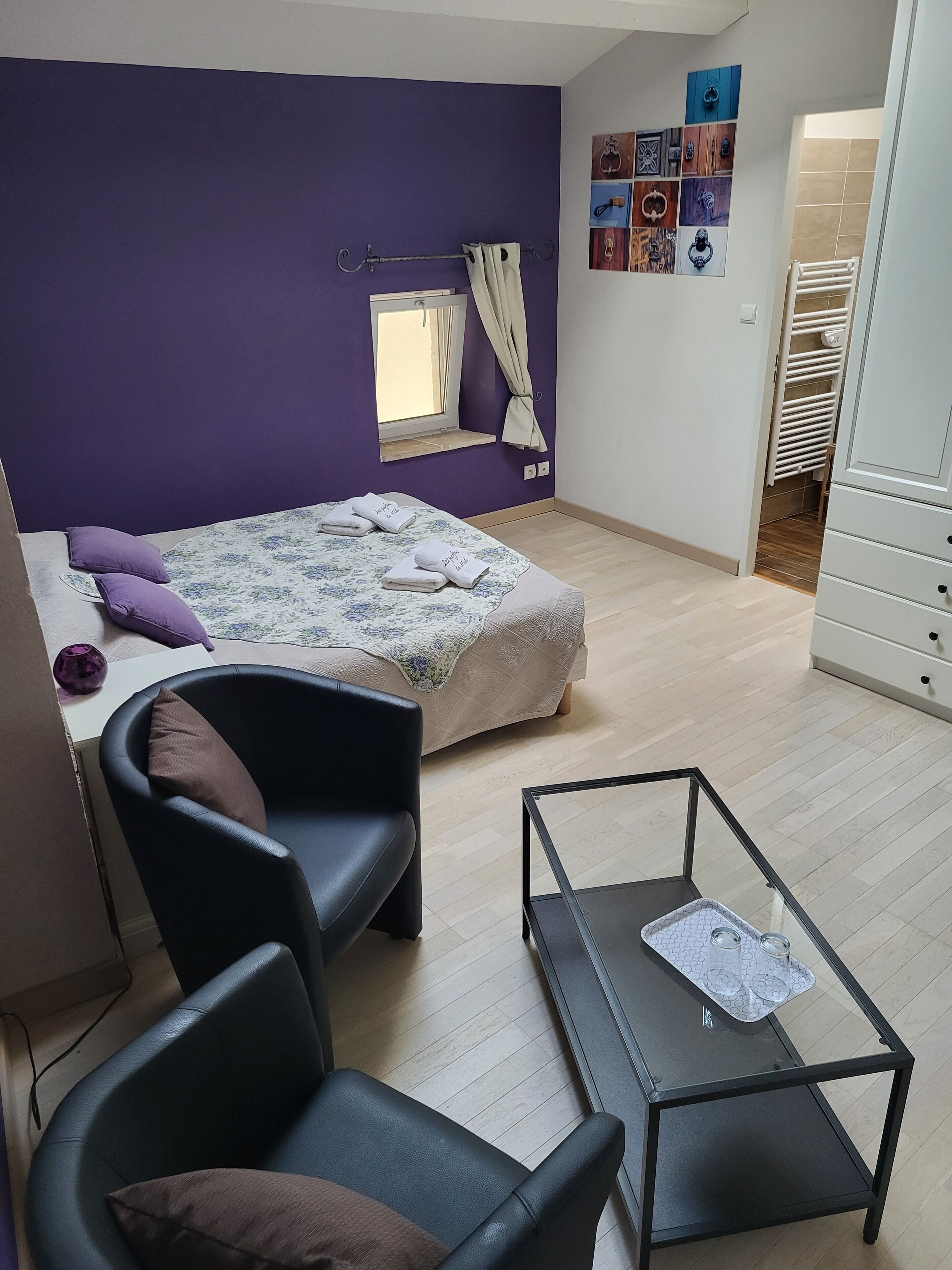 Un dormitorio compacto con cama, una silla, una mesita y un ventanal con cortinas.