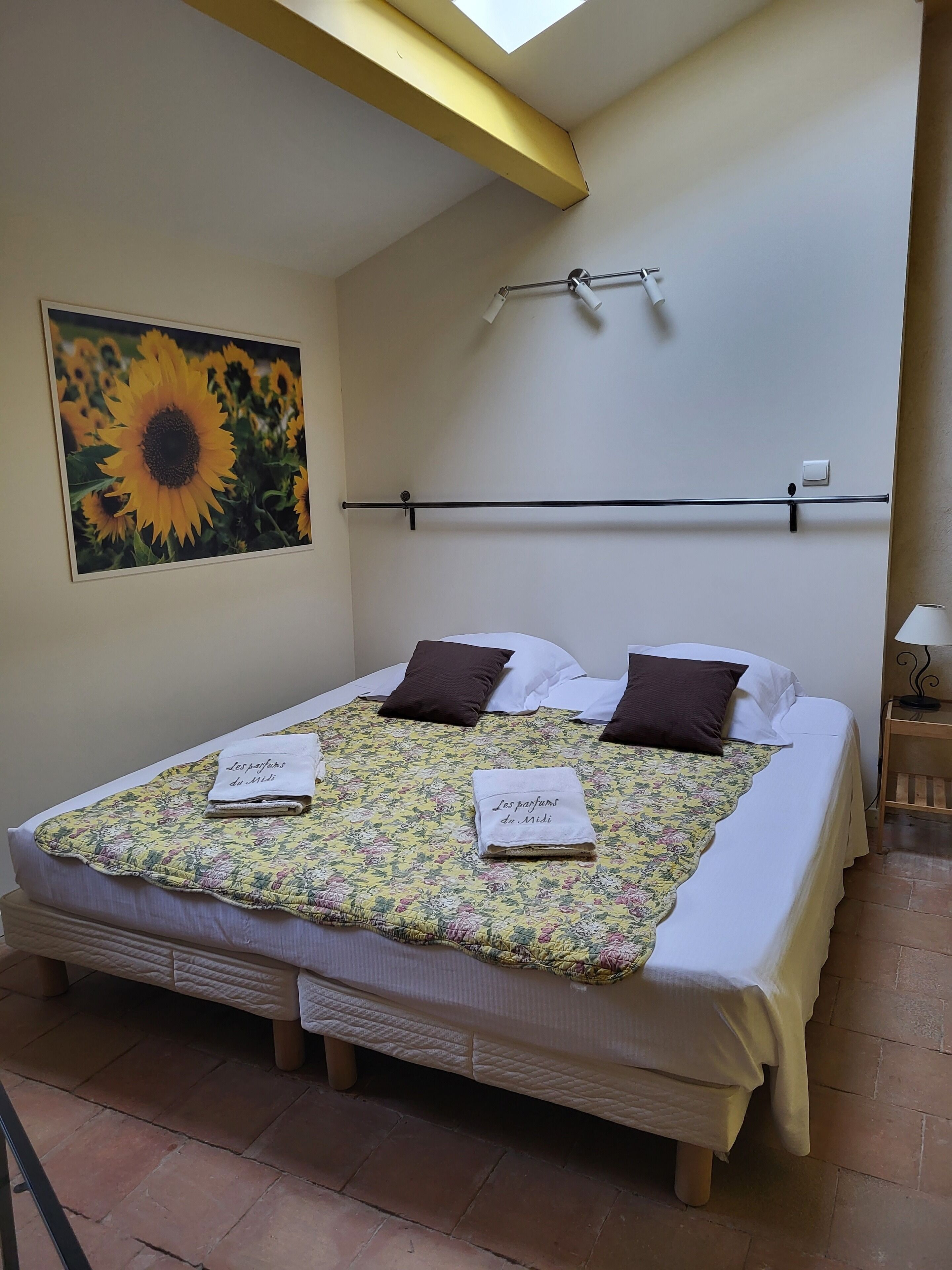 Una cama bien hecha con ropa de cama floral y dos toallas, una mesita de noche de madera con una lámpara, un cuadro de girasoles en la pared y un tragaluz arriba.