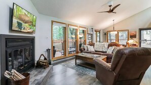 A cozy living room with a fireplace, comfortable seating, and large windows offering a view of the outdoors. - Rustic Retreat (Hedgesville)