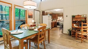 A spacious dining area with a wooden table and chairs, a hanging light fixture, and a view of the forest through large windows. - Rustic Retreat (Hedgesville)