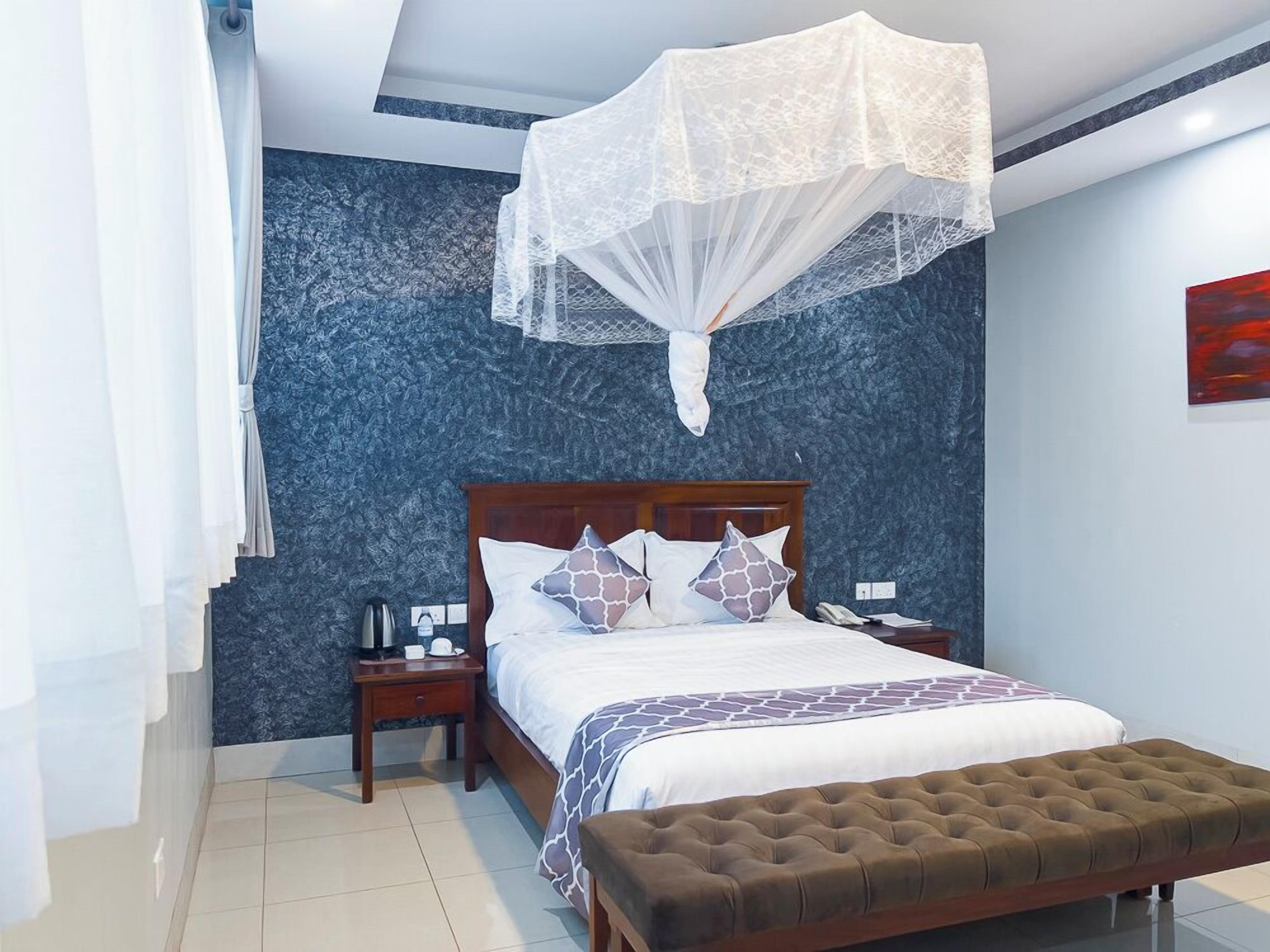 A bedroom with a bed, bedside tables, a nightstand, a bedside lamp, a window with curtains, and a wall-mounted artwork.