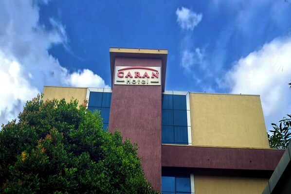 Un hotel llamado 'CARAN HOTEL' con un diseño arquitectónico moderno, que destaca un letrero prominente y un cielo azul despejado al fondo.