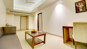 Un salón moderno con un sofá, una mesa de centro de madera y una silla. Hay un ventilador de techo, un cuadro enmarcado en la pared y una puerta que da a otra habitación.
