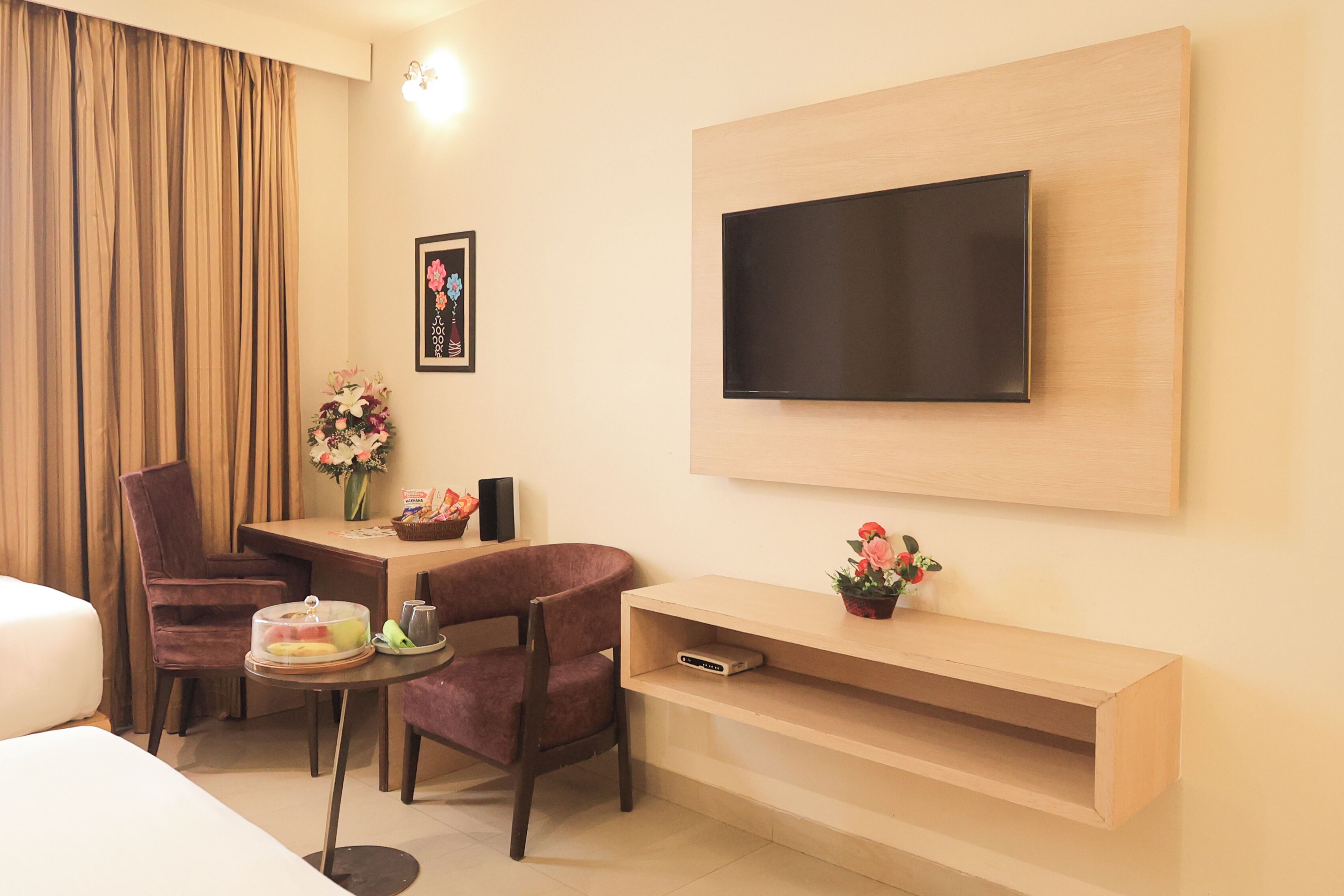 A hotel room with a bed, a desk with a chair, a TV mounted on the wall, and a small table with a vase of flowers.