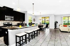 Una cocina moderna con una isla central, armarios negros y un microondas. Hay taburetes de bar, una zona de comedor con mesa y sillas, y un sofá en una sala de estar con ventanas amplias.