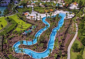 An aerial view of a resort with a winding pool, surrounded by palm trees and greenery.