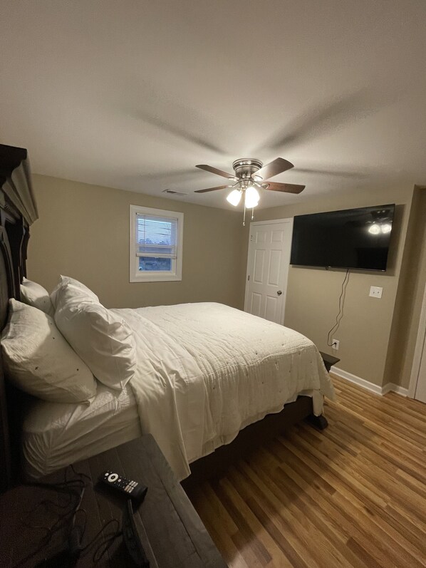 Un dormitorio con cama, ventilador de techo, televisor montado en la pared y ventana con persianas.