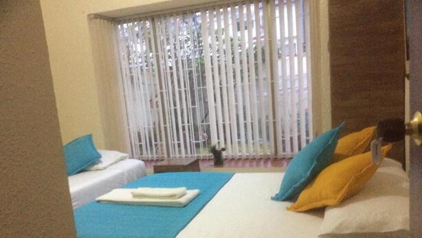 A hotel room with two beds, one with blue bedding and the other with white and yellow bedding. There is a window with vertical blinds, and a small table with a lamp in the corner. - The Palma Home (Bogotá)