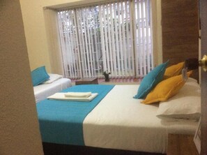 A hotel room with two beds, one with blue bedding and the other with white and yellow bedding. There is a window with vertical blinds, and a small table with a lamp in the corner. - The Palma Home (Bogotá)