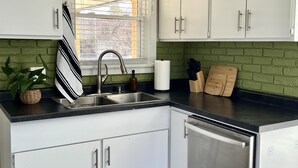 Une cuisine avec des armoires blanches, un lave-vaisselle en acier inoxydable, un évier avec un robinet, une fenêtre avec des stores, un pare-feu en briques vertes, un comptoir noir, une planche à découper en bois, un porte-couteaux et un tapis à motifs.