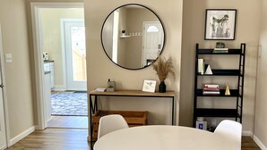 Un coin repas avec une table ronde et quatre chaises blanches, un ventilateur de plafond, un miroir et une bibliothèque contenant des livres et des objets décoratifs.