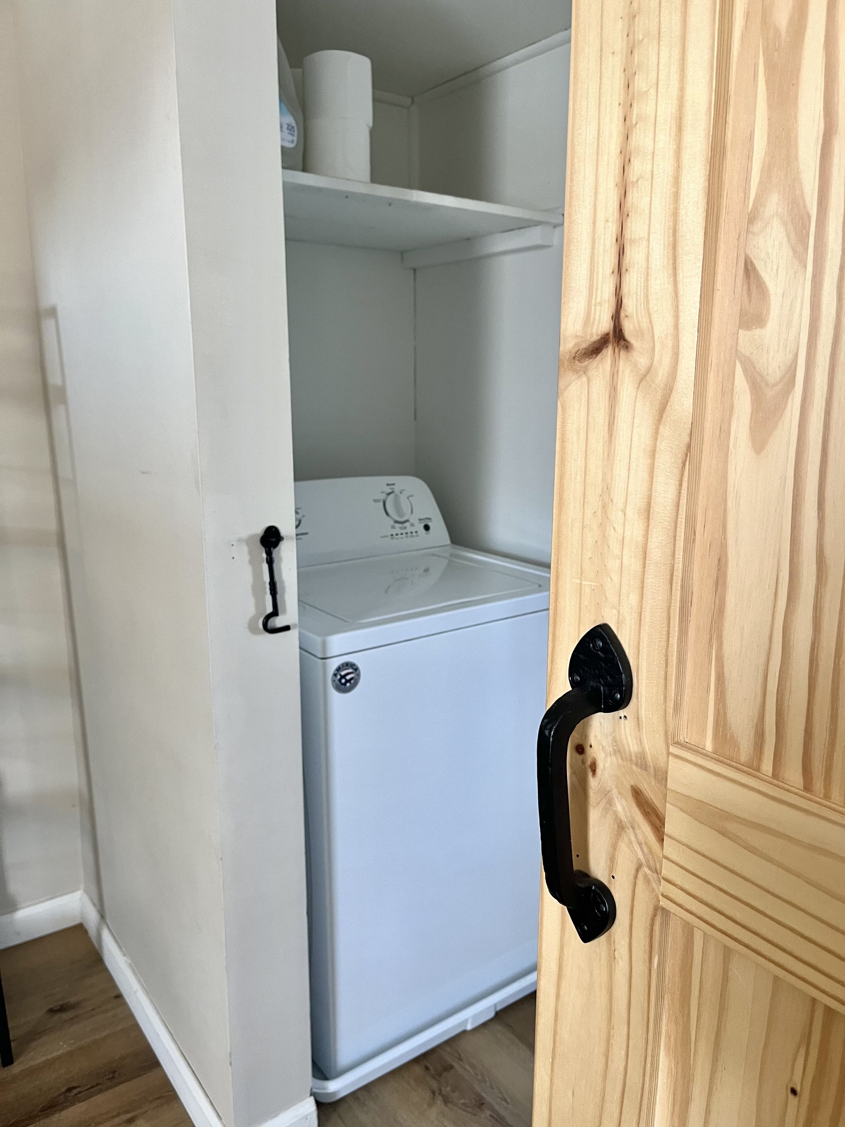 A compact, white laundry room with a built-in washer and dryer, a wooden door, and a shelf above the dryer.