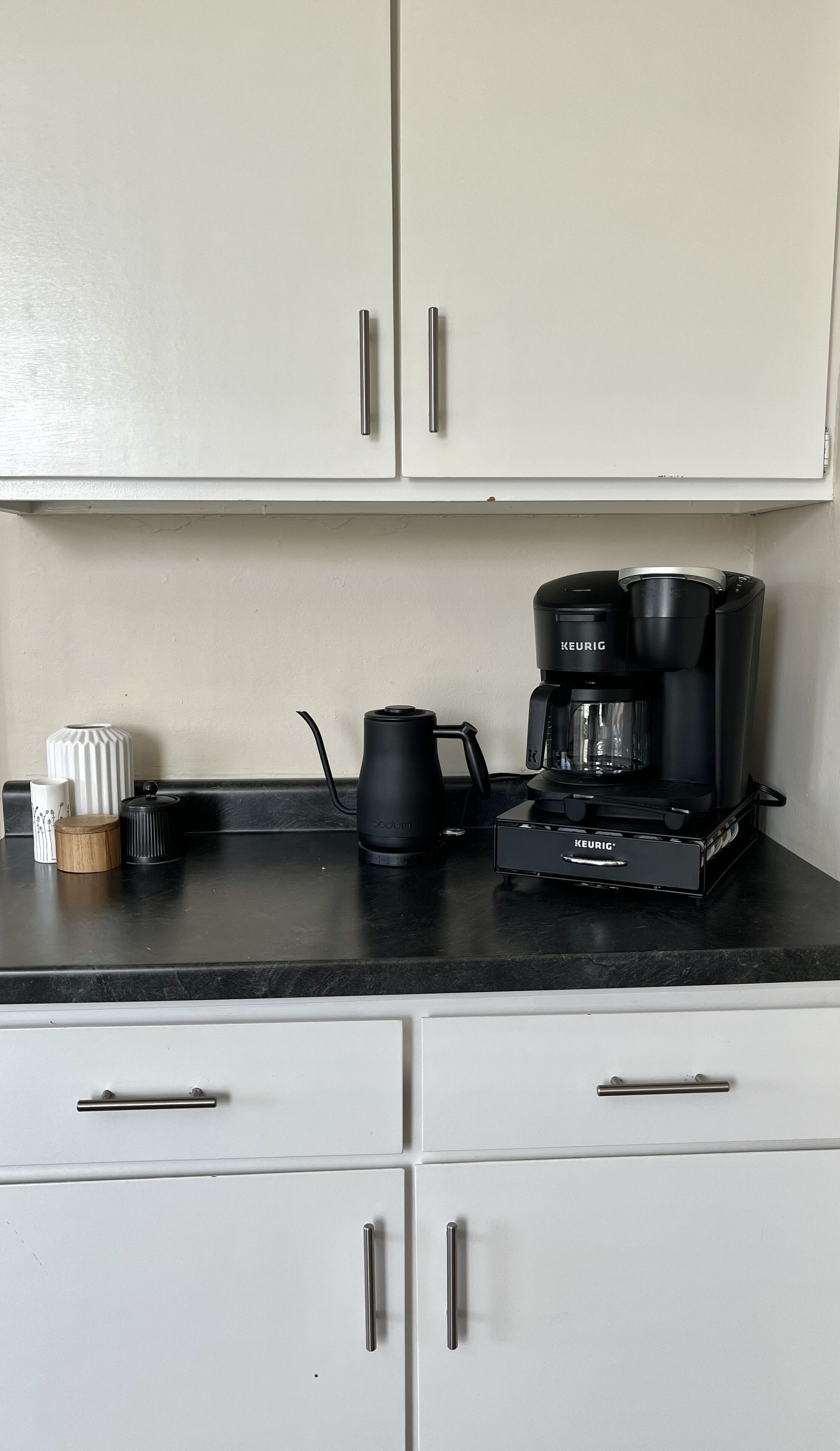 A kitchen with white cabinets, a black Keurig coffee maker, a Bodum coffee pot, and a small wooden container.