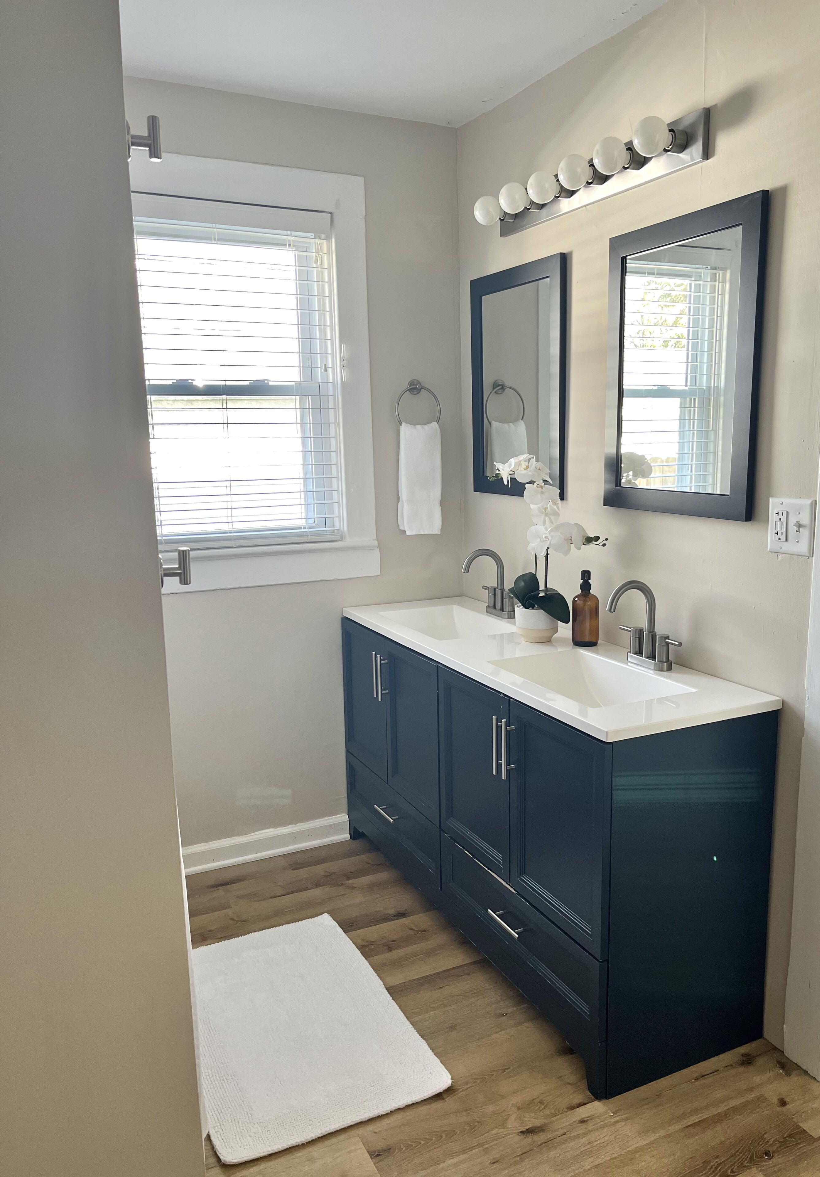 Une salle de bain avec un double lavabo, des meubles sombres, un plan de travail blanc, un tapis blanc et une fenêtre avec des stores.