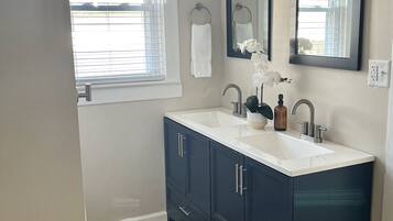 Une salle de bain avec un double lavabo, des meubles sombres, un plan de travail blanc, un tapis blanc et une fenêtre avec des stores.