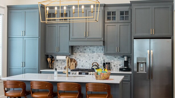 Een moderne keuken met grijze kastjes, een centraal eiland met barkrukken, een roestvrijstalen koelkast en een hanglamp.