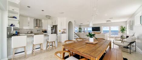 Una cocina moderna con armarios blancos, una mesa de comedor de madera y vista al océano.