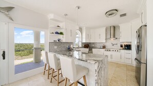 Une cuisine moderne avec des meubles blancs, un îlot en marbre doté de tabourets, des appareils en acier inoxydable et une vue sur la verdure environnante.