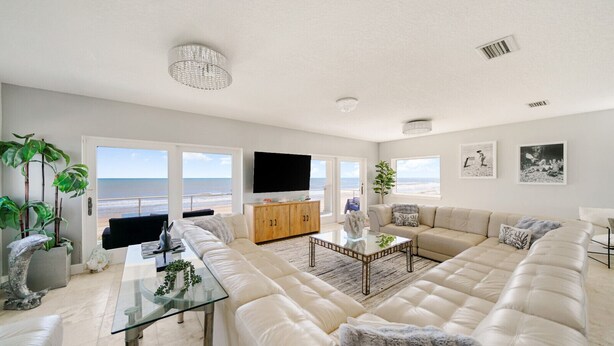 Un salon spacieux avec un grand canapé d’angle beige, une table basse en verre et un meuble en bois. De grandes fenêtres offrent une vue sur la plage.