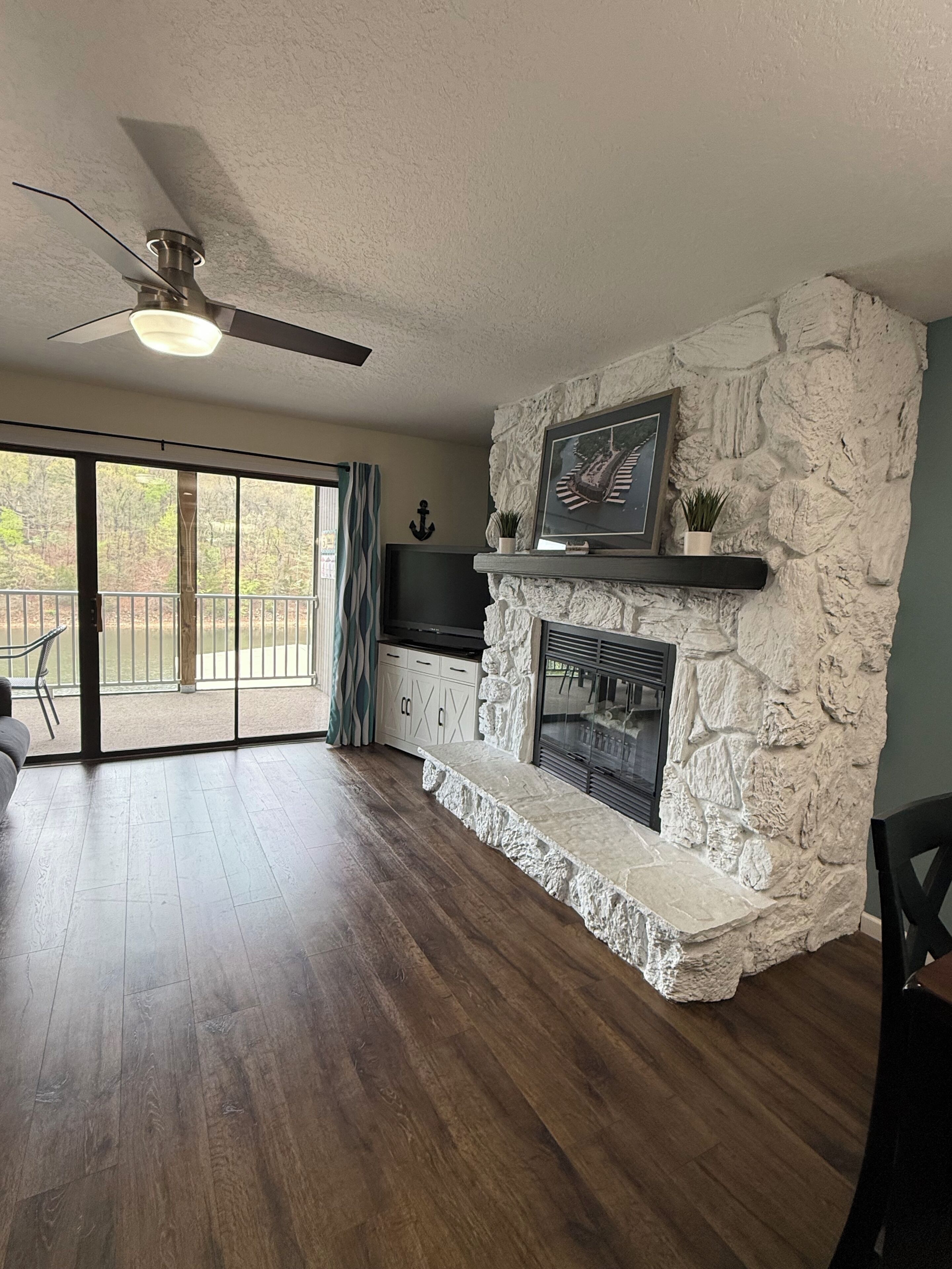 A living room with a stone fireplace, a ceiling fan, and a view through large glass doors.