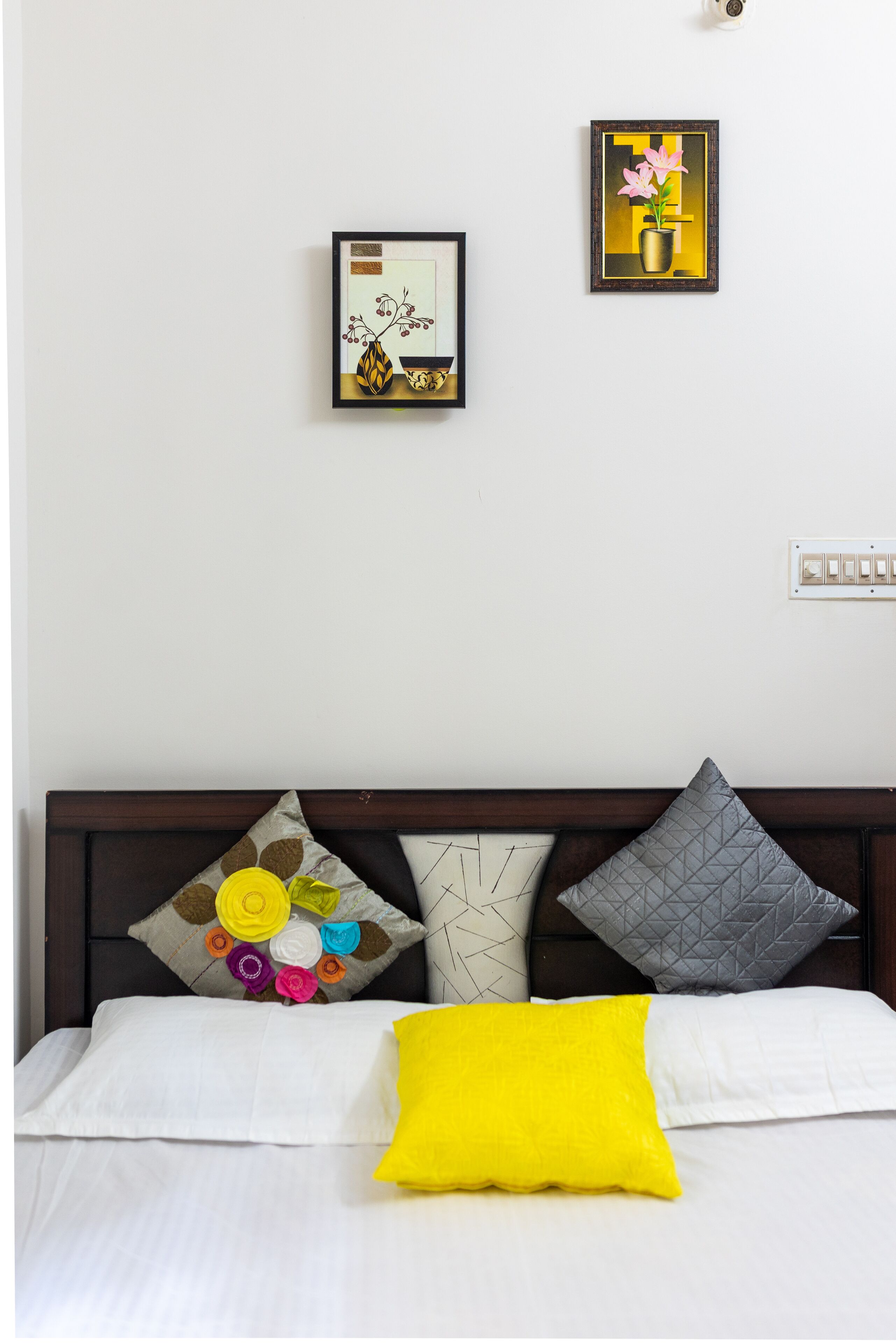 A neatly made bed with white bedding and a bright yellow pillow. Above the bed, there are two framed artworks on the wall.