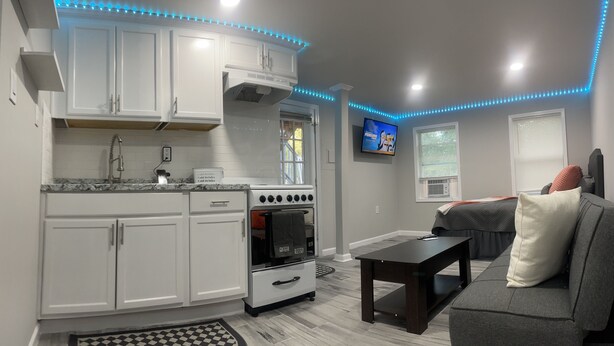 Una cocina con gabinetes blancos, una estufa y un lavamanos. Hay un televisor en la pared y una ventana con persianas.