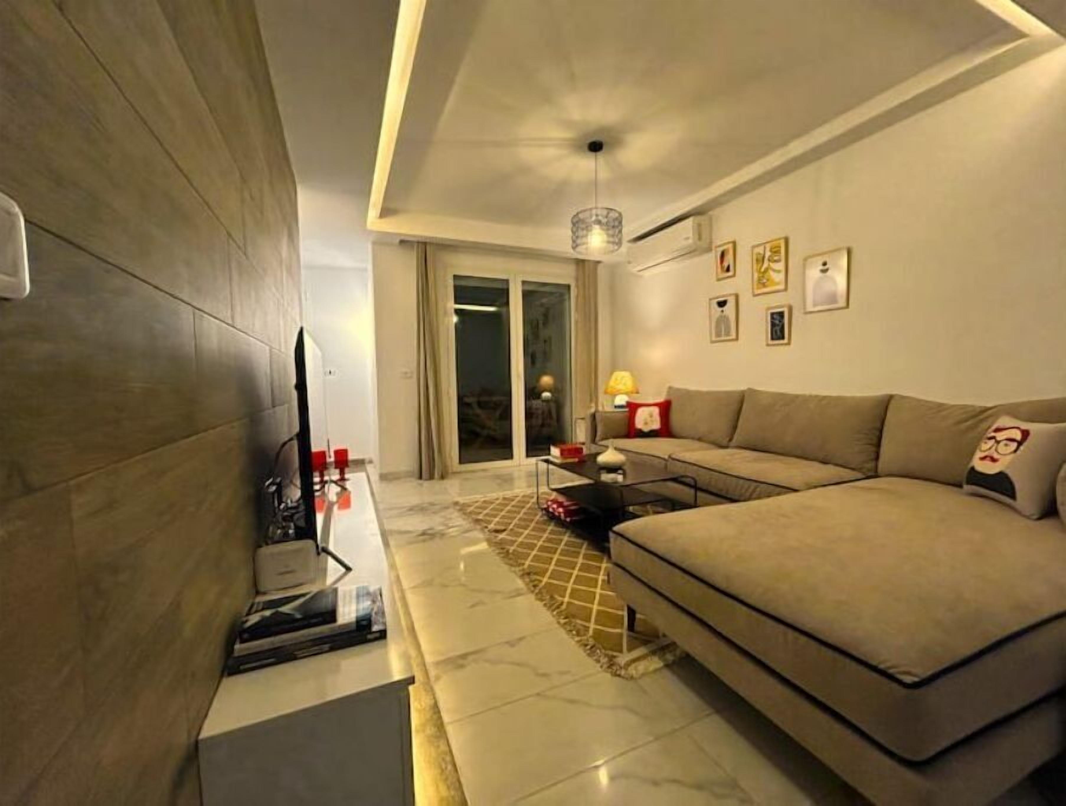 A modern living room with a sofa, a coffee table, and a TV. There is a sliding glass door leading to a balcony.