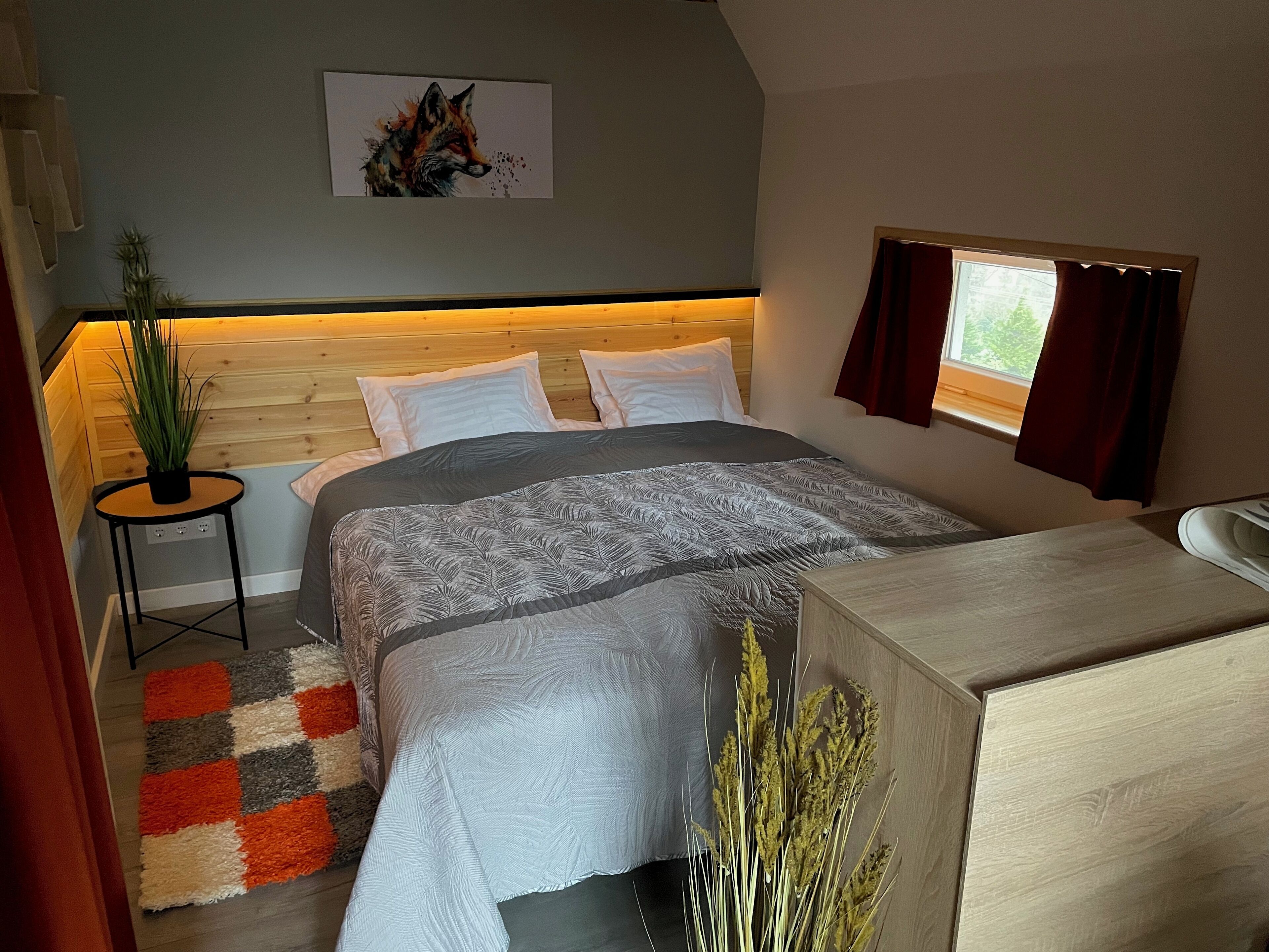 Un dormitorio con cama, mesita de noche, planta en maceta, ventana con cortinas y un cuadro en la pared.