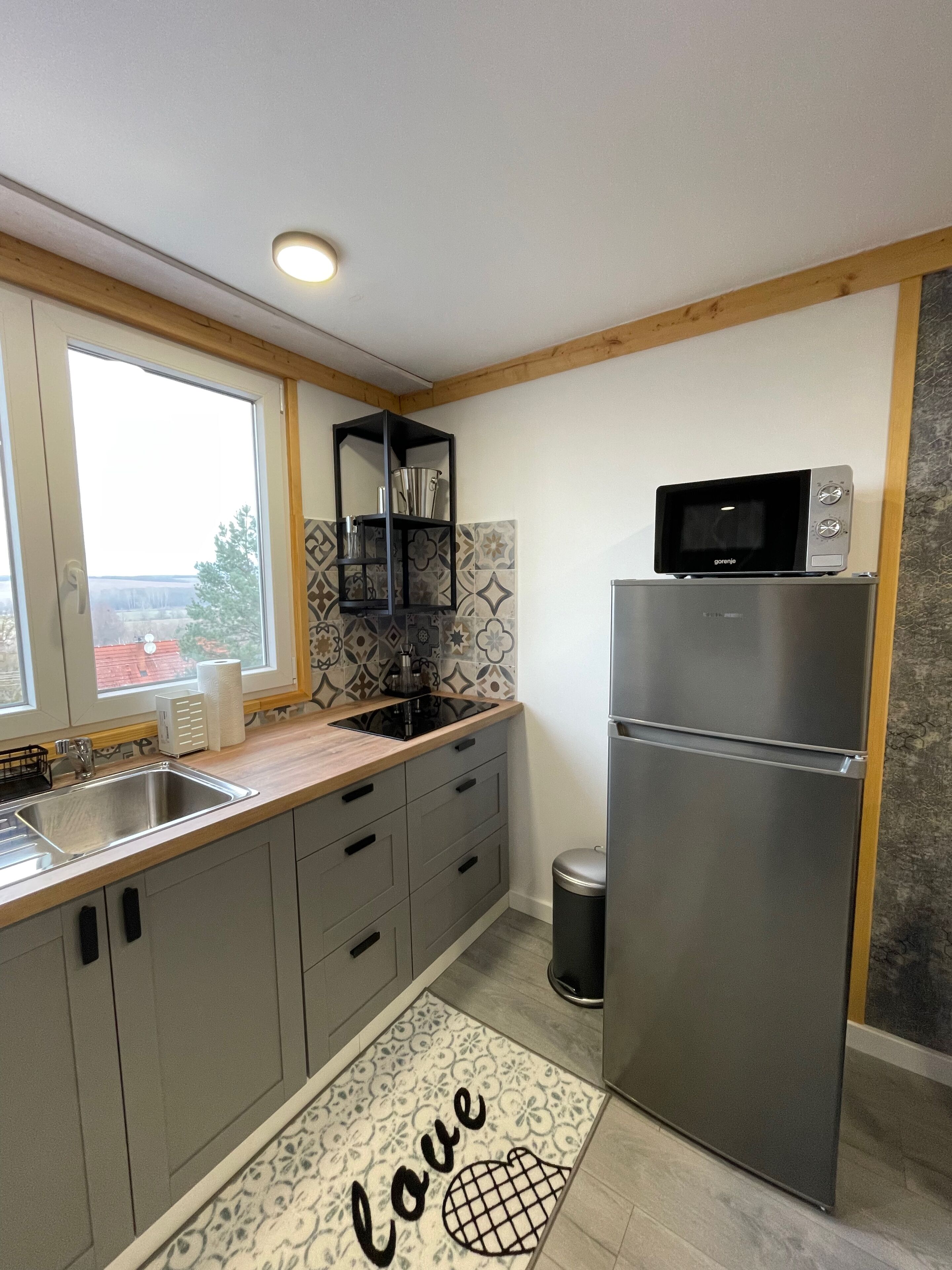 A modern kitchen with a stainless steel refrigerator, a microwave, and a built-in oven. There is a window providing a view of the outside, and a shelf unit above the sink.