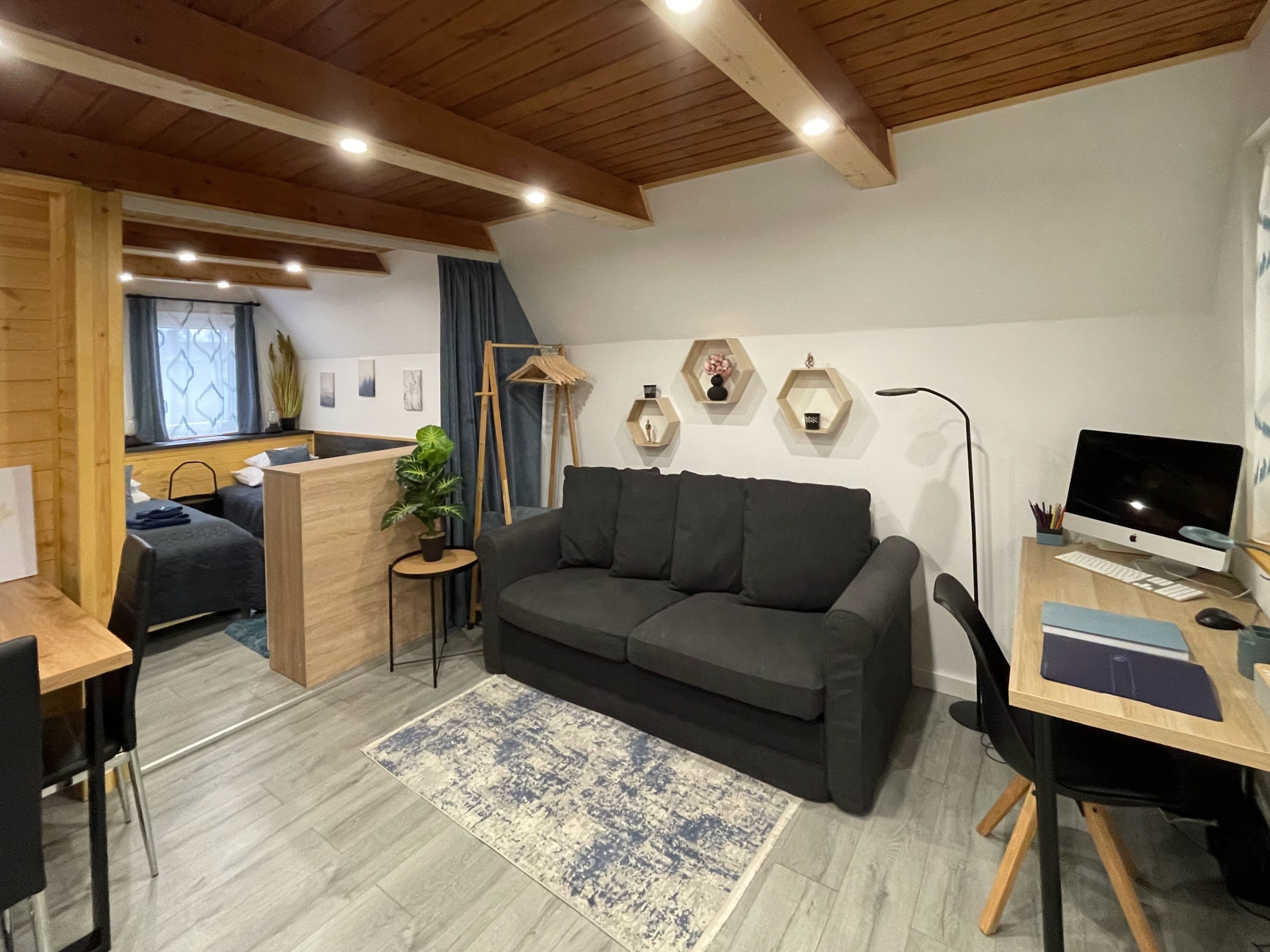 A modern living room with a grey sofa, wooden ceiling beams, a desk with a computer, and a small side table with a plant.