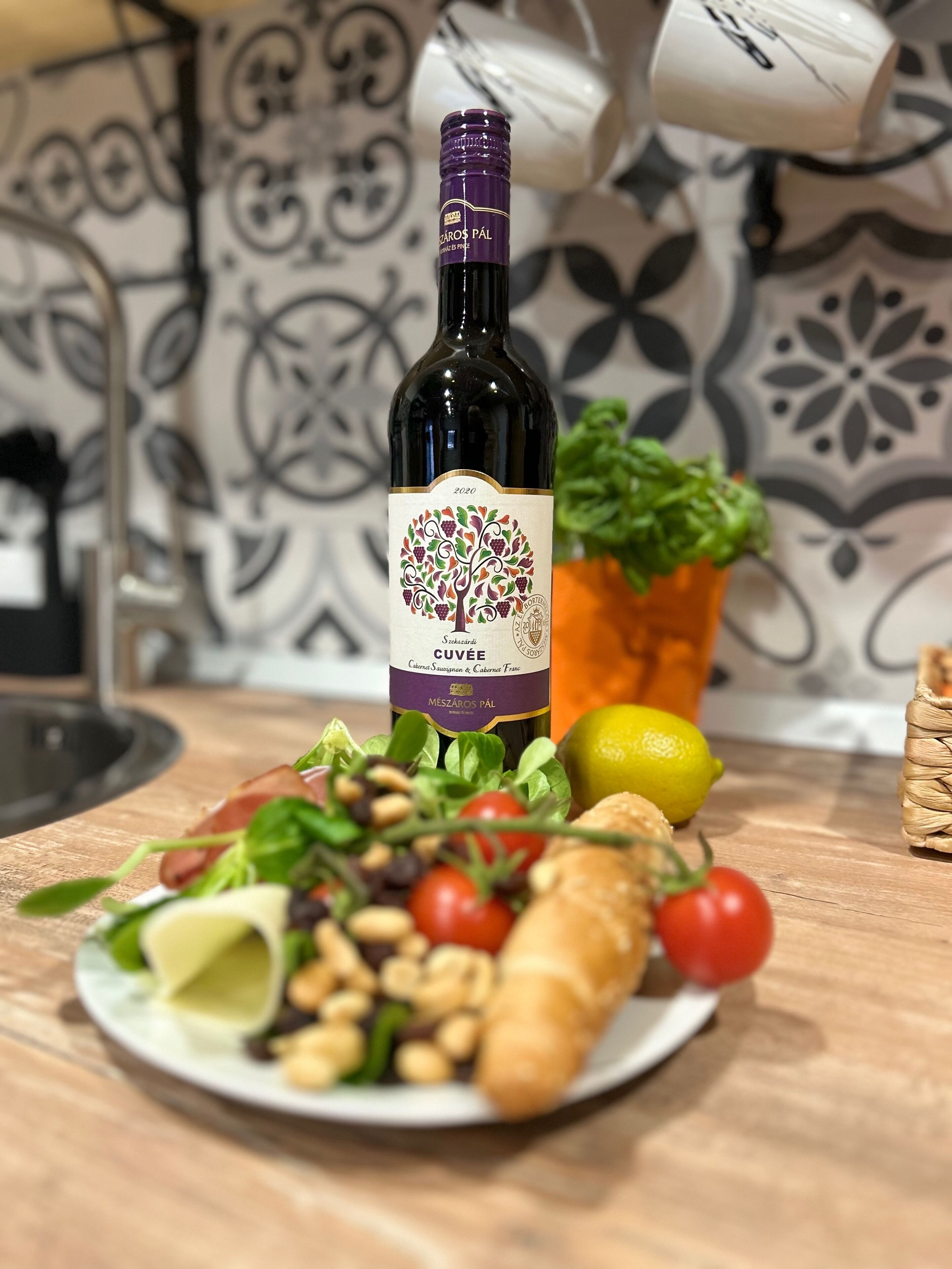 Una botella de vino con etiqueta que muestra un árbol y uvas, colocada sobre una mesa con un plato de comida que incluye pan, tomates y verduras.