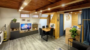 A modern, well-lit room with a wooden ceiling, a TV on a shelf, a dining table with chairs, and a small desk area. - Calm Resort Hertelend (Magyarhertelend)