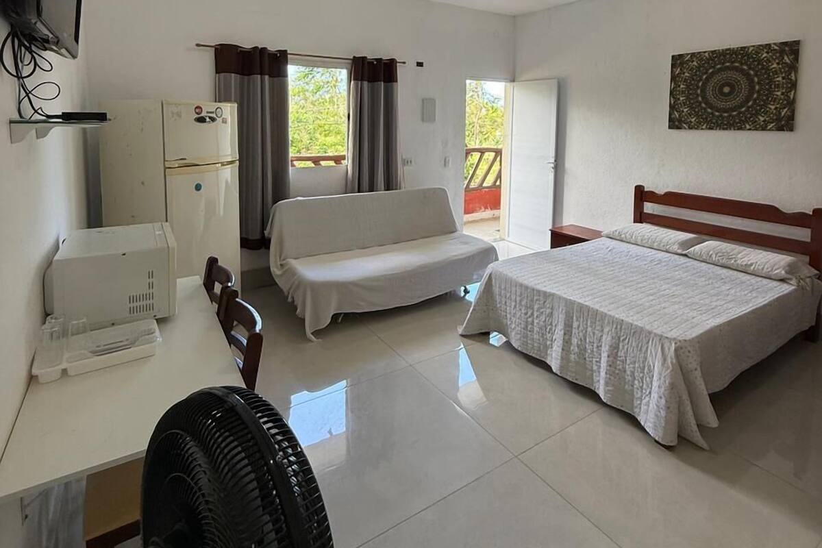 A hotel room with a bed, a sofa, a desk with a computer, a fan, and a TV.