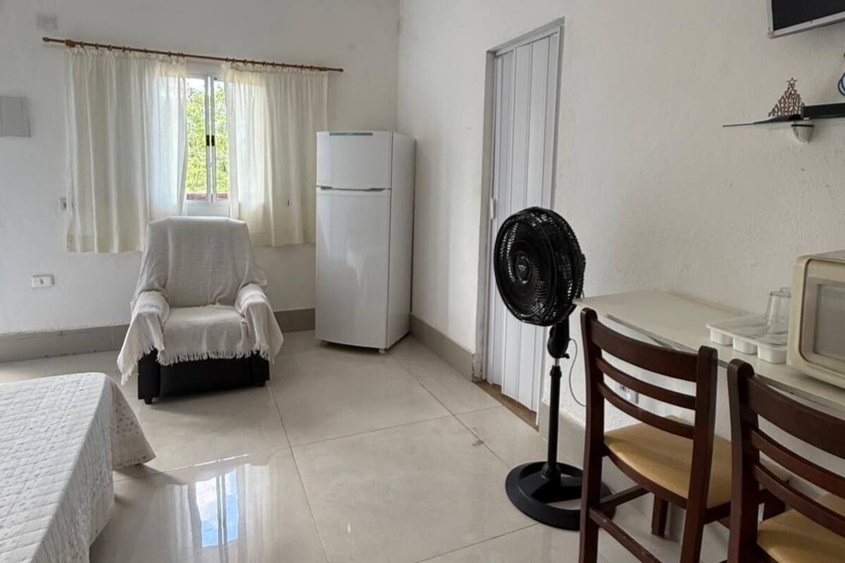 A room with a bed, a chair, a table, a fan, a refrigerator, and a television.