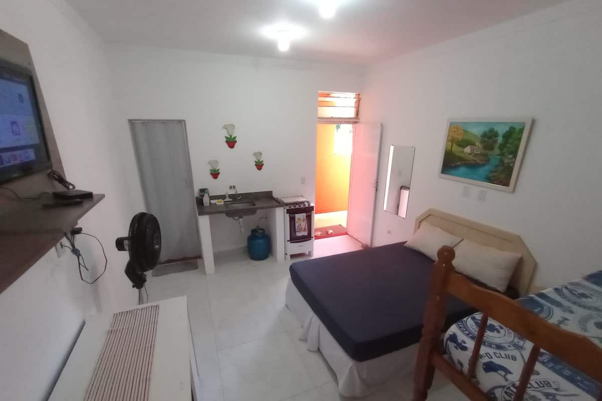 A compact room with a bed, a desk with a chair, a small sink, and a fan.