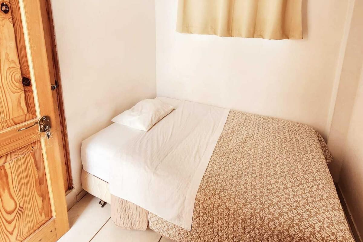 Una habitación pequeña con cama individual, puerta de madera, ventana y cortina.