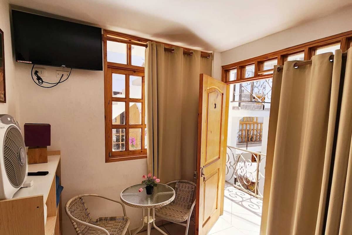 Una habitación pequeña con televisión, ventilador, mesa con sillas y ventana con cortinas.