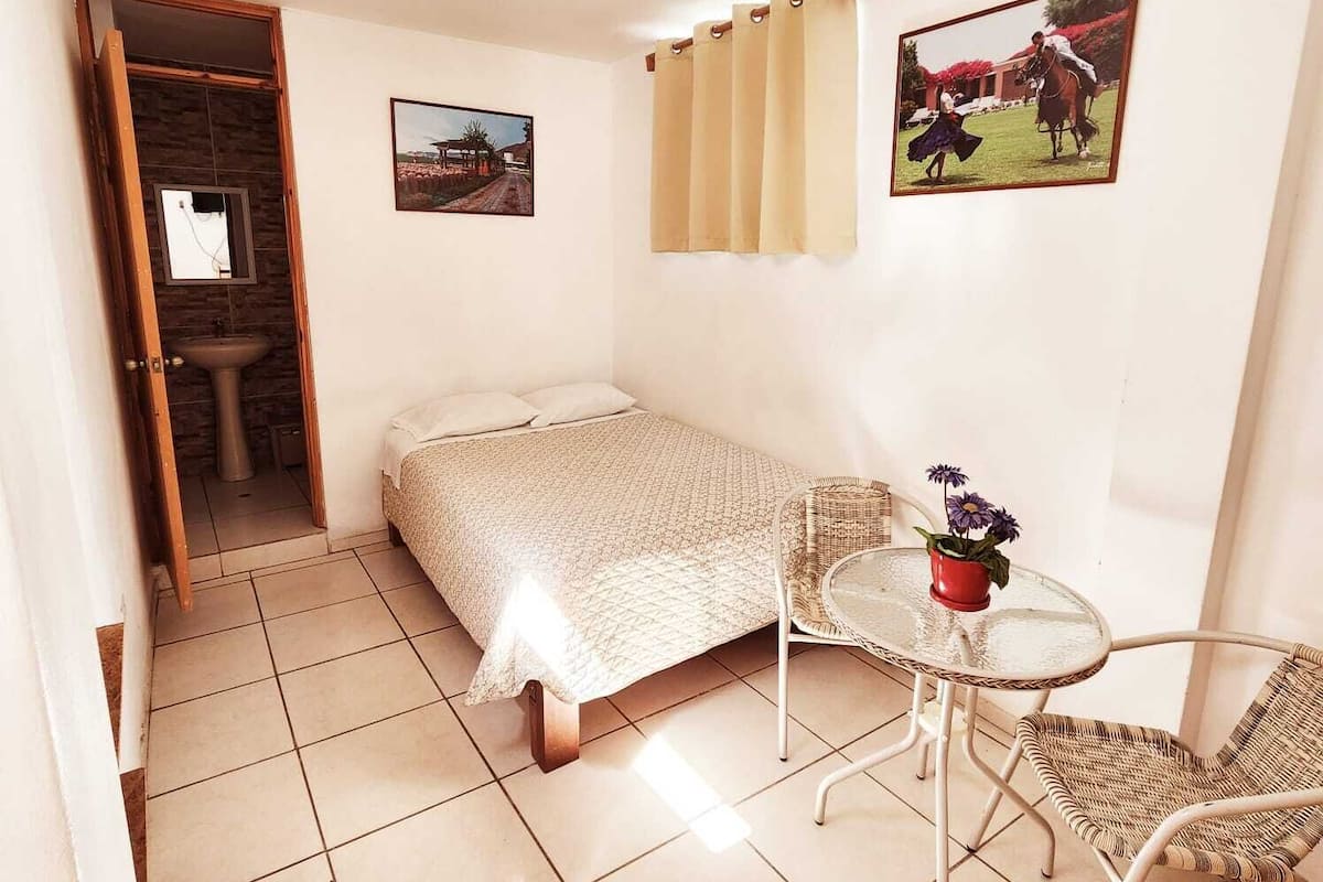 Una habitación pequeña y bien iluminada con una cama, una mesita con una silla, un lavamanos y dos cuadros en la pared.